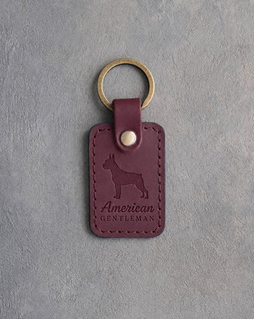 Sangria red leather keychain debossed with a Boston Terrier silhouette and the words American GENTLEMAN, on gray concrete