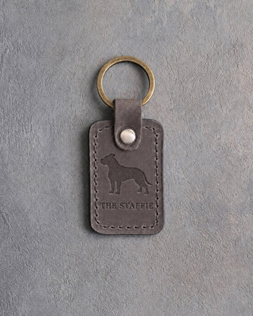 Country gray leather keychain debossed with an American Staffordshire Terrier silhouette above THE STAFFIE, on gray concrete