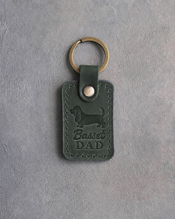 Forest green leather keychain debossed with a Basset Hound silhouette and the words Basset DAD, on gray concrete