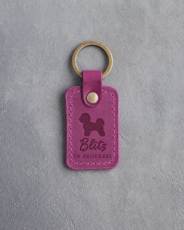 Fuchsia pink leather keychain debossed with a Bichon Frise and the words Blitz IN PROGRESS, on gray concrete