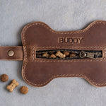 Brown bone shaped leather treat pouch stamped BUDDY, zipper slightly open with treats peeking out