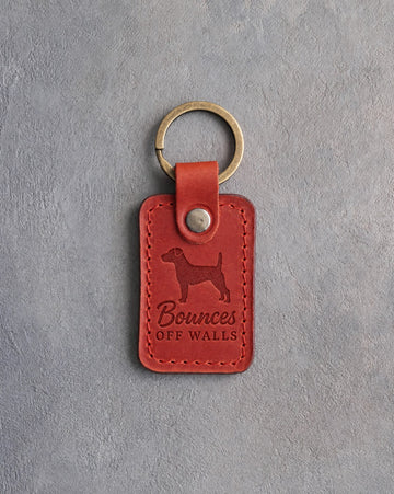 Poppy red leather keychain debossed with a Jack Russell terrier and the words Bounces OFF WALLS, on gray concrete