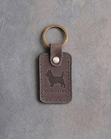 Country gray leather keychain debossed with a Cairn Terrier silhouette above THE CAIRN, on gray concrete