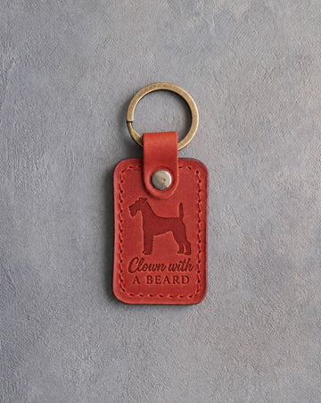 Poppy red leather keychain debossed with an Airedale terrier and the words Clown with A BEARD, on gray concrete
