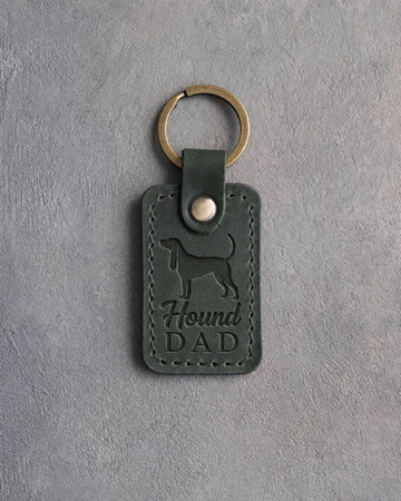 Forest green leather keychain debossed with a Coonhound silhouette and the words Hound DAD, on gray concrete