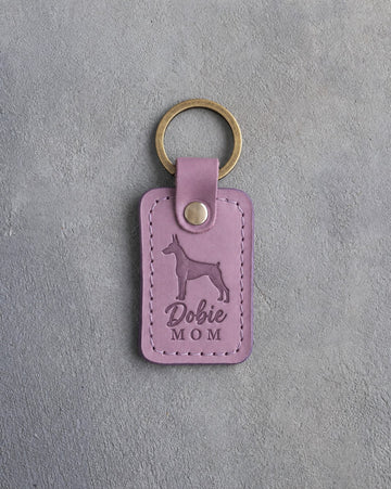 Lilac leather keychain debossed with a Doberman silhouette and the words Dobie MOM, on gray concrete