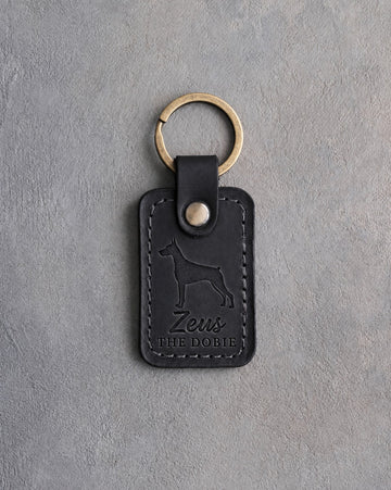 Black leather keychain debossed with a Doberman silhouette, the name Zeus and THE DOBIE, on gray concrete