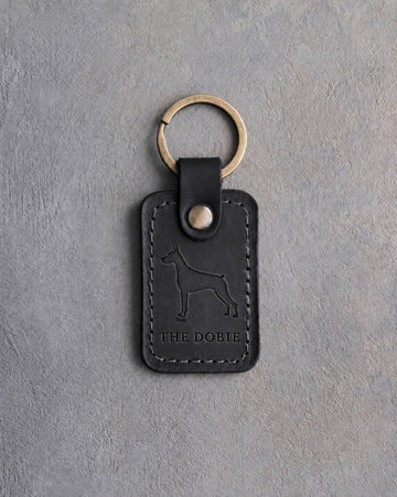 Black leather keychain debossed with a Doberman silhouette above THE DOBIE, on gray concrete