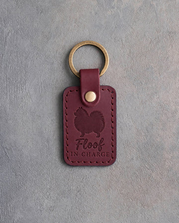 Sangria red leather keychain debossed with a Pomeranian silhouette and the words Floof IN CHARGE, on gray concrete