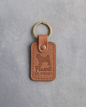 Caramel leather keychain debossed with a pug silhouette and the words Fluent in SNORT, on gray concrete