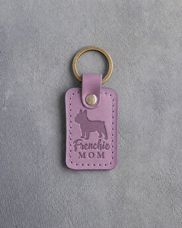 Lilac leather keychain debossed with a French bulldog silhouette and the words Frenchie MOM on gray concrete