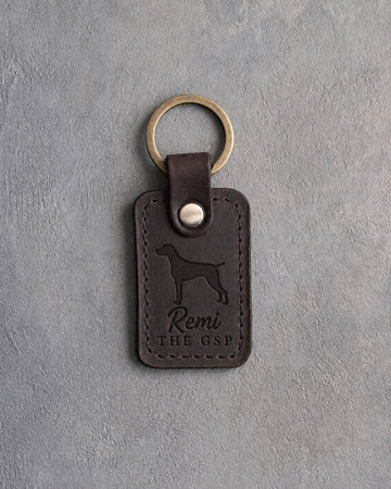 Dark espresso leather keychain debossed with a German Shorthaired Pointer silhouette, the name Remi and THE GSP, on gray concrete