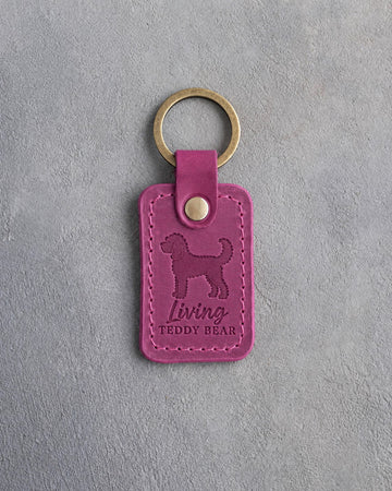 Fuchsia pink leather keychain debossed with a curly goldendoodle and the words Living TEDDY BEAR, on gray concrete