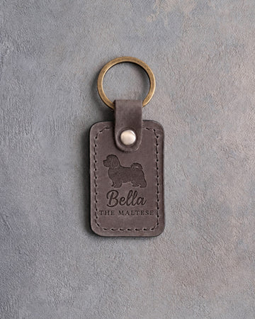 Country gray leather keychain debossed with a Maltese silhouette, the name Bella and THE MALTESE, on gray concrete