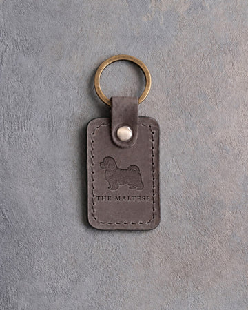Country gray leather keychain debossed with a Maltese silhouette above THE MALTESE, on gray concrete