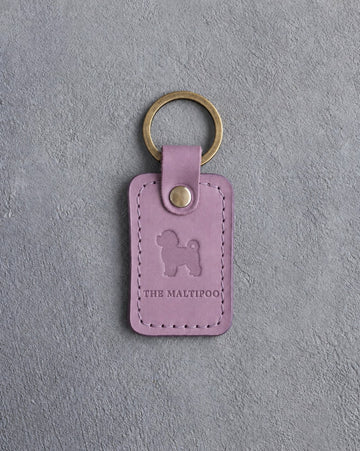 Lilac leather keychain debossed with a tiny maltipoo above THE MALTIPOO, on gray concrete