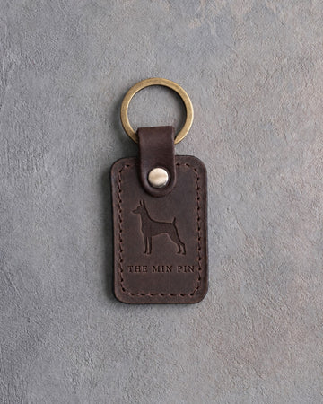 Dark espresso leather keychain debossed with a miniature pinscher above THE MIN PIN, on gray concrete