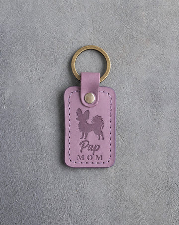 Lilac leather keychain debossed with a Papillon silhouette and the words Pap MOM, on gray concrete
