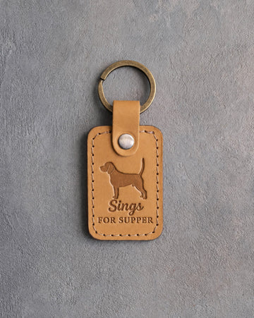 Indian Summer tan leather keychain debossed with a beagle silhouette and the words Sings FOR SUPPER, on gray concrete