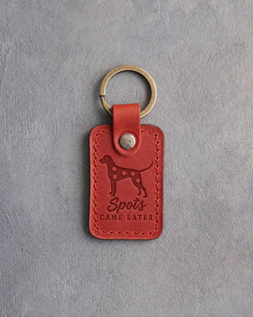 Poppy red leather keychain debossed with a Dalmatian silhouette and the words Spots CAME LATER, on gray concrete