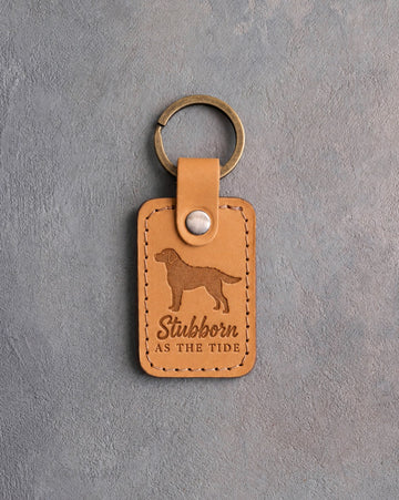 Indian Summer golden leather keychain debossed with a Chesapeake Bay retriever and the words Stubborn AS THE TIDE, on gray concrete