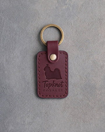 Sangria red leather keychain debossed with a Shih Tzu silhouette and the words Topknot ROYALTY, on gray concrete