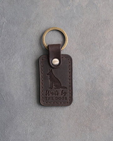 Dark espresso leather keychain debossed with a German shepherd sitting at attention and the words Waits by THE DOOR, on gray concrete