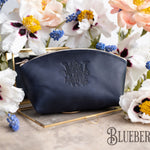 Leather makeup bag with a custom initial in blueberry navy, floral monogram on a gold tray amid white blooms