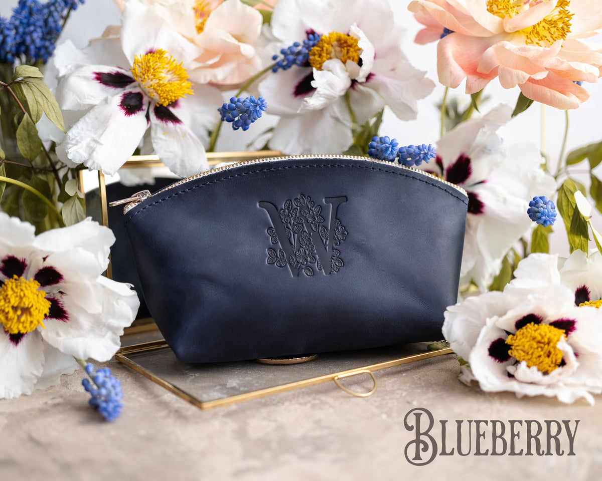 Leather makeup bag with a custom initial in blueberry navy, floral monogram on a gold tray amid white blooms