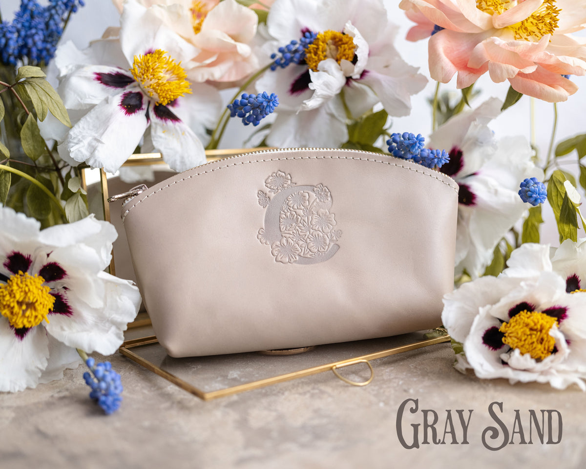 Gray sand leather makeup pouch with a floral monogram deboss, styled on a gold tray with fresh flowers