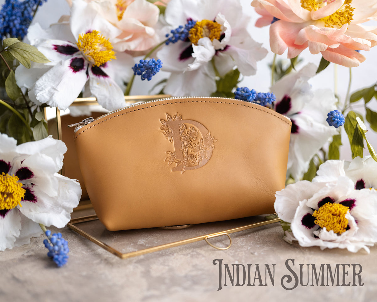 Indian summer golden-tan leather makeup bag with an ornate monogram, white flowers behind