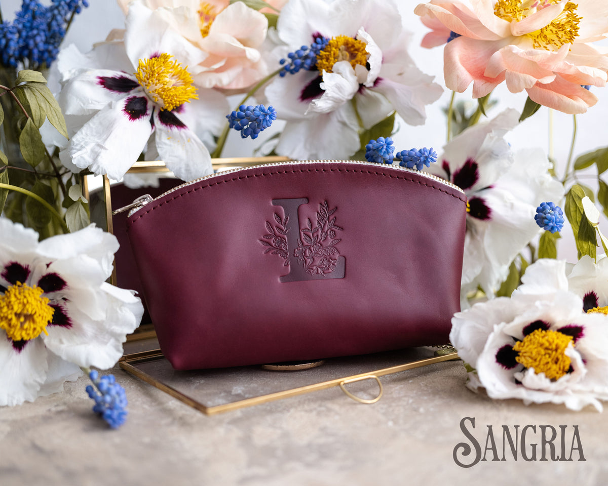 Sangria burgundy leather makeup pouch with a floral initial L, muscari and blooms behind