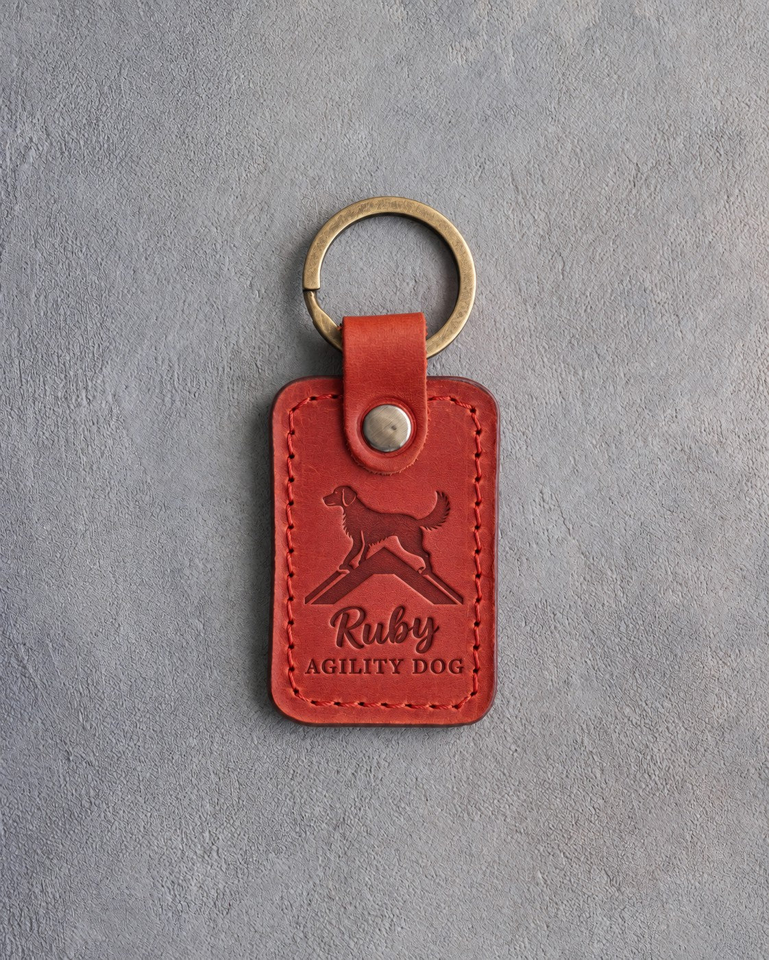 Poppy red leather keychain debossed with a dog on top of an agility A-frame above Ruby and AGILITY DOG, on gray concrete