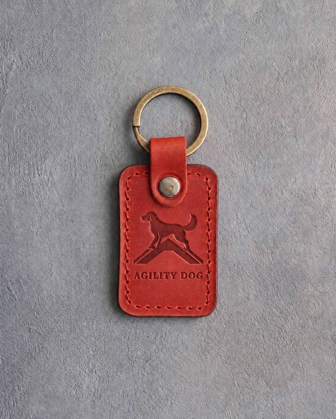 Poppy red leather keychain debossed with a dog on top of an agility A-frame above AGILITY DOG, on gray concrete