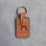 Caramel leather keychain debossed with a bearded Airedale terrier above THE AIREDALE, on gray concrete