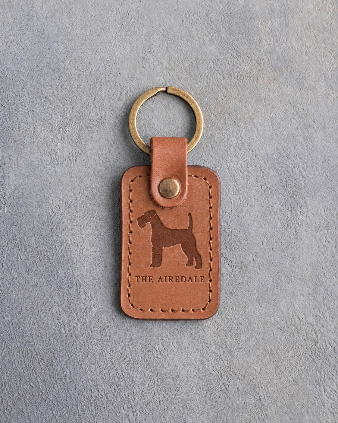 Caramel leather keychain debossed with a bearded Airedale terrier above THE AIREDALE, on gray concrete