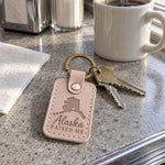 Alaska Raised Me keychain in pale gray leather clipped to a parka pocket on a frosty morning