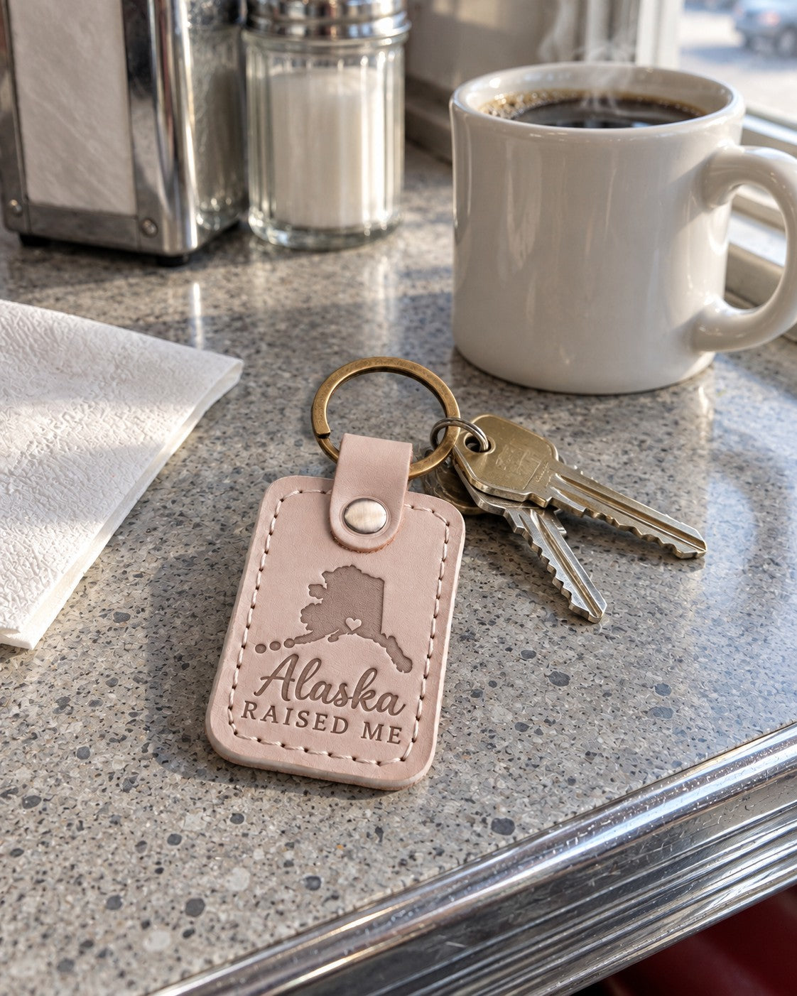 Alaska Raised Me keychain in pale gray leather clipped to a parka pocket on a frosty morning