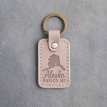Gray sand leather keychain debossed with an Alaska silhouette and a heart at Anchorage above Alaska and RAISED ME, on gray concrete