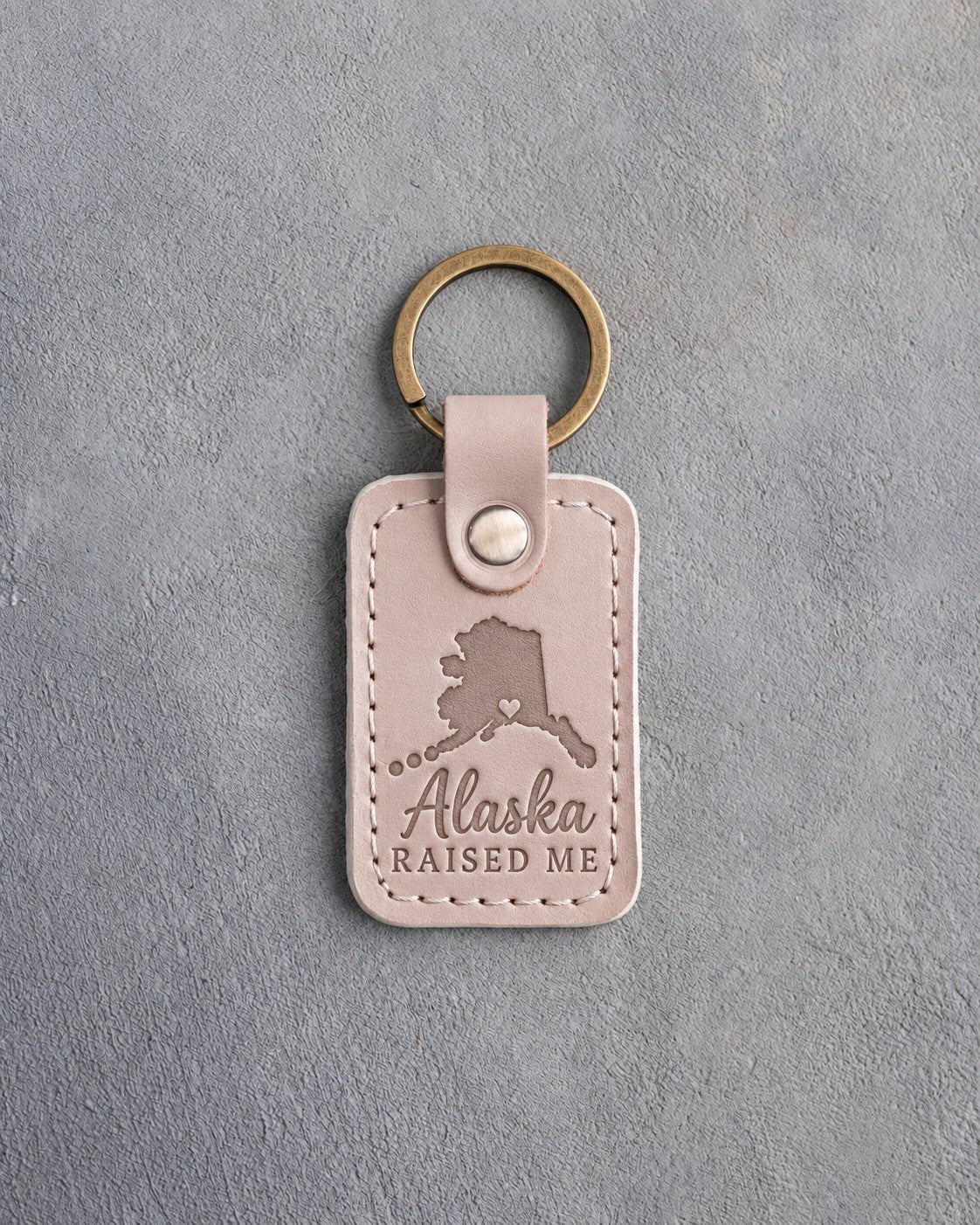 Gray sand leather keychain debossed with an Alaska silhouette and a heart at Anchorage above Alaska and RAISED ME, on gray concrete