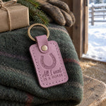 Lilac All I want IS A HORSE keychain on a folded green wool blanket inside a barn door, a fir bough behind it and snow on the corral rails