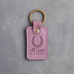 Lilac leather keychain debossed with a heels-up horseshoe above All I want and IS A HORSE, on gray plaster