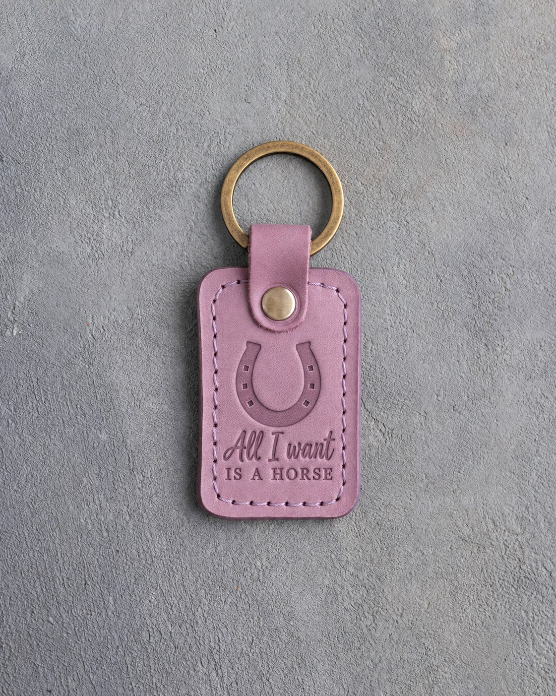 Lilac leather keychain debossed with a heels-up horseshoe above All I want and IS A HORSE, on gray plaster