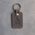 Country gray leather keychain debossed with a small seated mixed breed dog on a horizon line above Always BY MY SIDE, on gray concrete