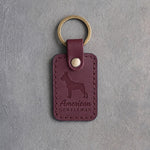 Sangria red leather keychain debossed with a Boston Terrier silhouette and the words American GENTLEMAN, on gray concrete