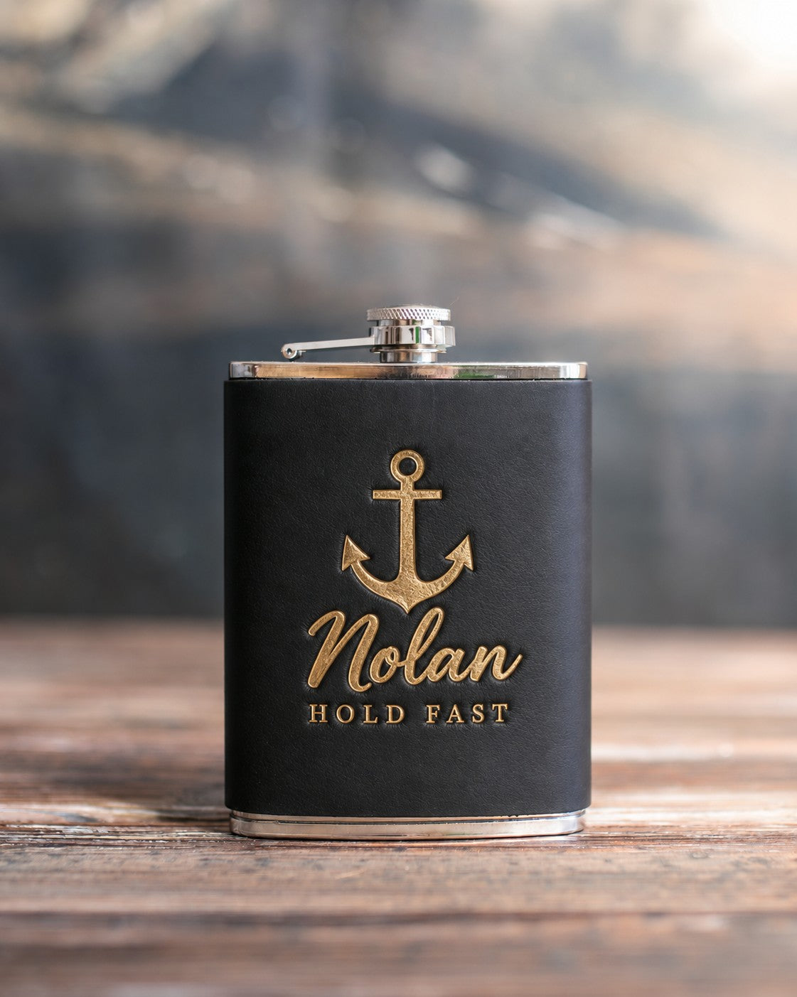 Black leather-wrapped hip flask stamped in gold foil with an admiralty anchor above Nolan and HOLD FAST, on a wooden table