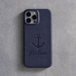 Back of a blueberry leather iPhone case debossed with an admiralty anchor above Nolan and HOLD FAST below the camera, on gray plaster