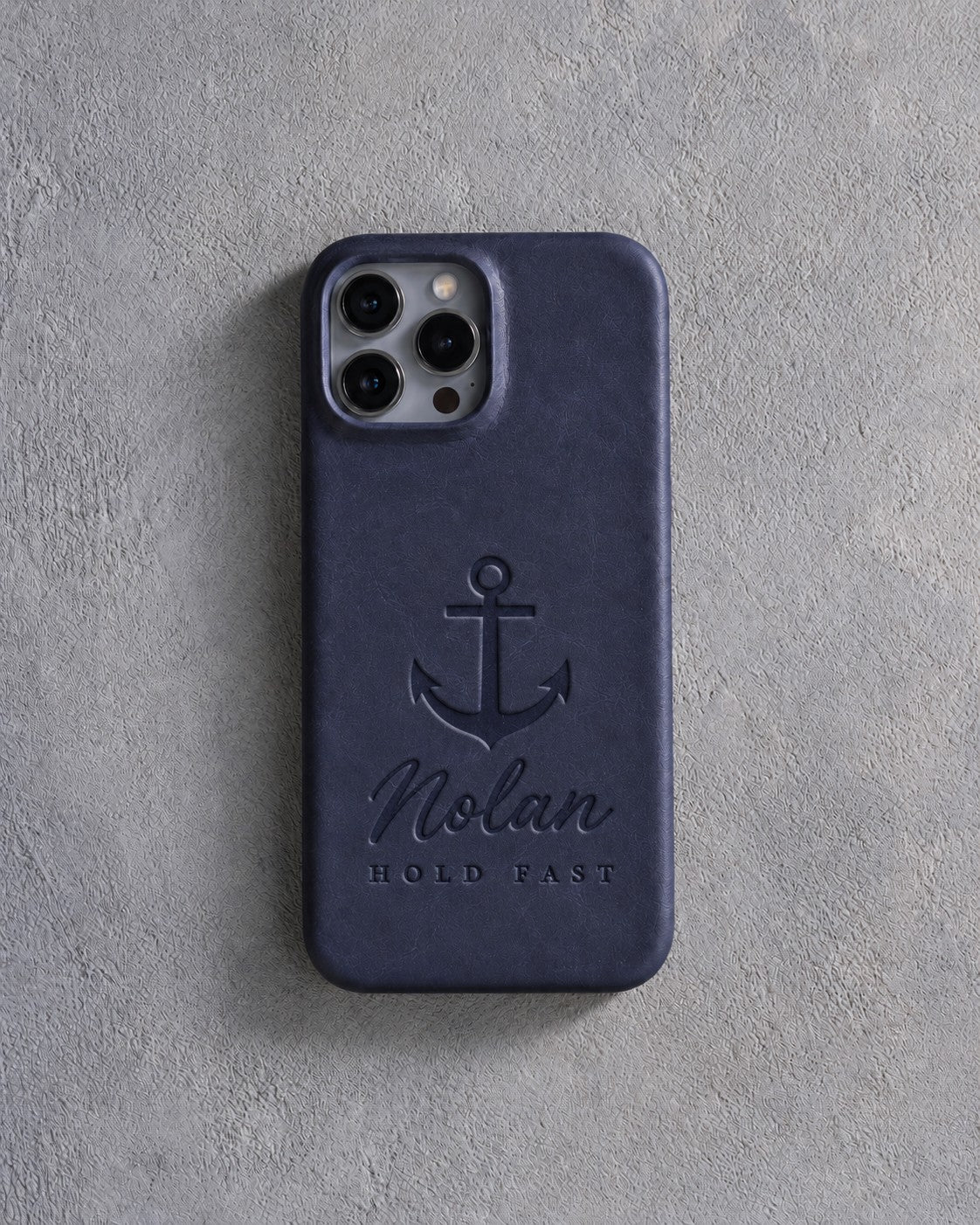 Back of a blueberry leather iPhone case debossed with an admiralty anchor above Nolan and HOLD FAST below the camera, on gray plaster