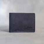 Closed blueberry leather bifold wallet with an admiralty anchor beside Nolan and HOLD FAST pressed in the lower right corner, on gray plaster
