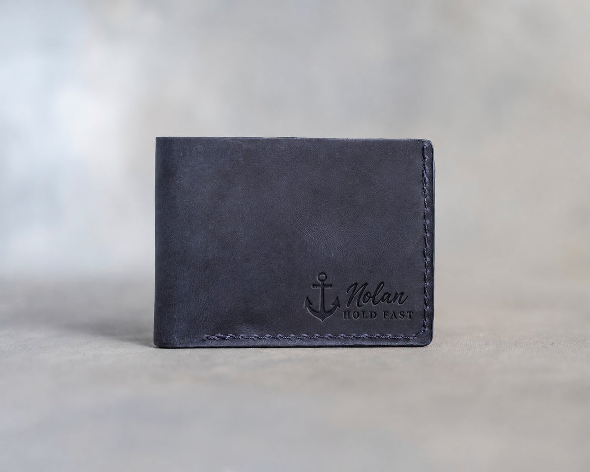 Closed blueberry leather bifold wallet with an admiralty anchor beside Nolan and HOLD FAST pressed in the lower right corner, on gray plaster